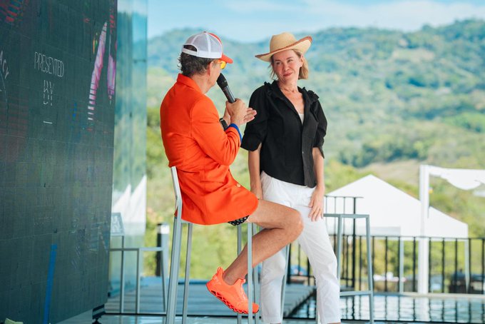 A man in an orange blazer and cap holds a microphone while interviewing a woman in a black shirt and straw hat on an outdoor stage.