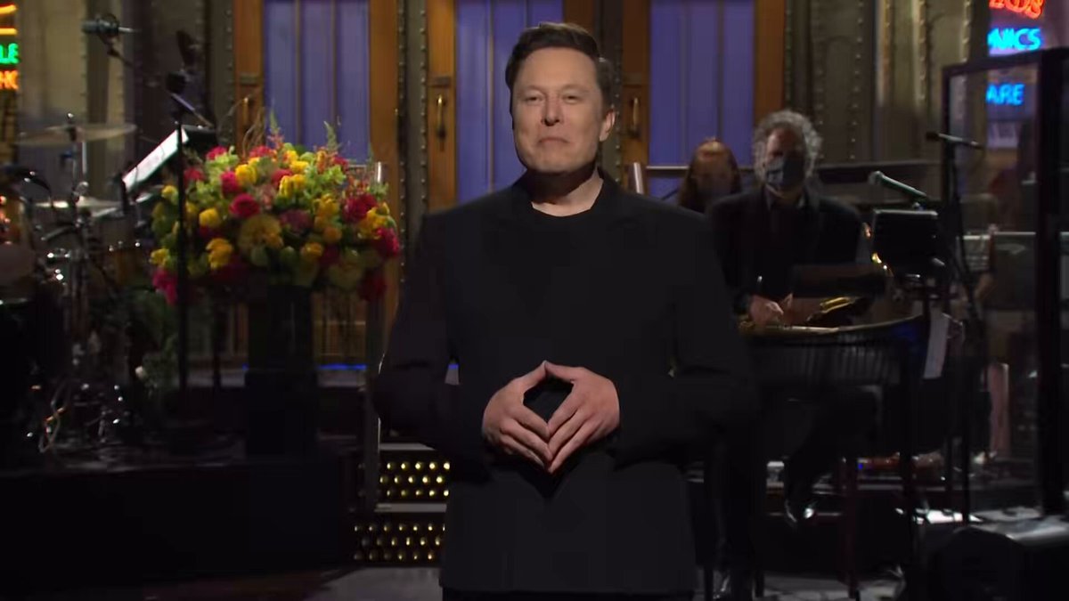 The image features a man standing in front of a stage, dressed in a black suit with his hands clasped together. The background includes a stage set up for a performance or event, as well as some flowers placed nearby. There are also other people present in the scene, but they are not the main focus of the image.

The post that accompanies this image is titled "":- @elonmusk was born 69 days after 4/20. He must be an avatar of someone's video game." The post seems to be a humorous or lighthearted commentary on the man in the suit, possibly referencing his character from a video game.