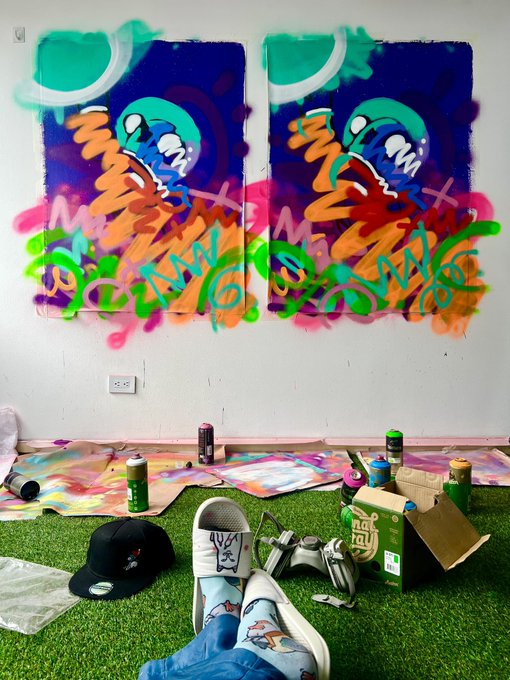 A photo showing two vibrant spray-painted artworks on a wall depicting monster-like faces, with the artist's perspective featuring feet in sneakers, a hat, spray paint cans, and paint-covered cardboard on artificial grass below.