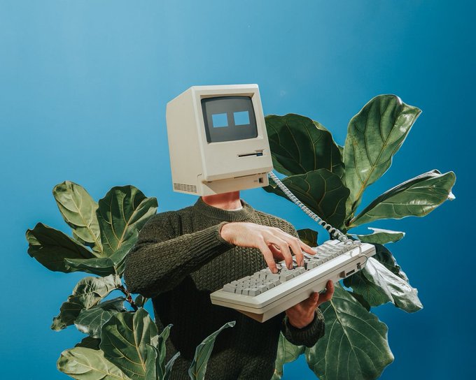 A person wearing a vintage Macintosh computer as a head and a green sweater types on a retro keyboard, surrounded by large green leaves against a blue background.