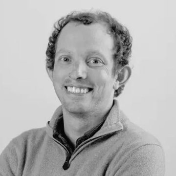 The image features a man with curly hair wearing a gray sweater. He has a big smile on his face as he looks directly at the camera. The photo appears to be black and white, giving it an artistic touch. There is no text present in the image, but there is a caption that reads "Just In: Nathan Allman, founder of Ondo finance passes away." This information seems to be related to the man's death, as he is described as being "passed" by someone else.