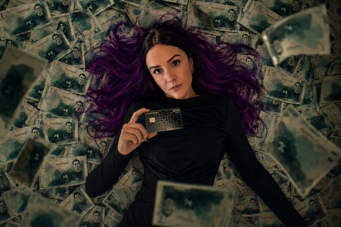 A woman with vibrant purple hair lies surrounded by scattered hundred-dollar bills while holding up a new Visa credit card, visually symbolizing wealth and excitement. The image complements the post's enthusiasm about receiving a "new badge" and proudly representing @hyperbeat on Hyperliquid, with the cash and card reinforcing themes of financial success and crypto earnings.