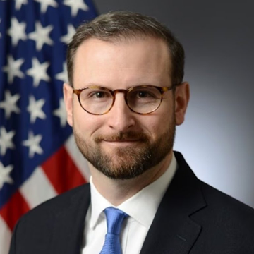 An official portrait of a man with glasses and a beard wearing a suit and blue tie, standing in front of an American flag.