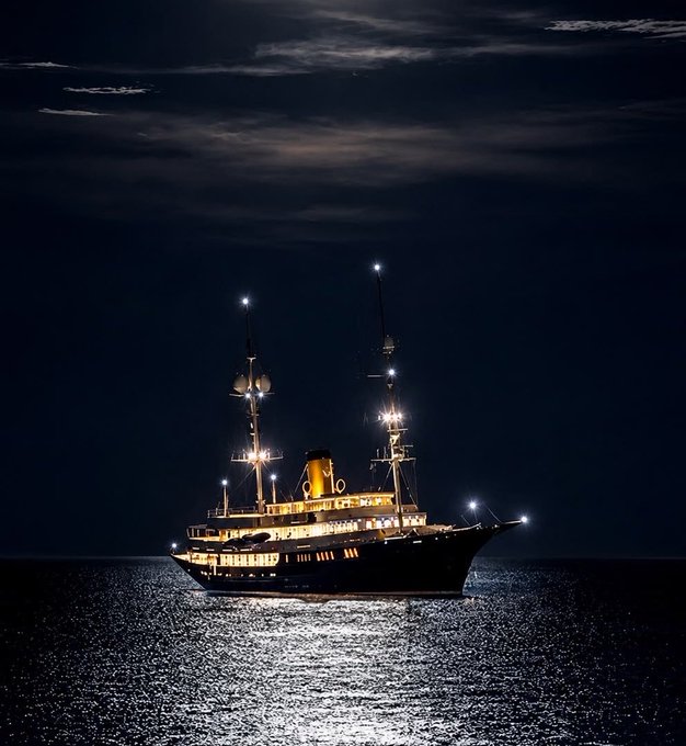 A large illuminated multi-masted ship sailing on the water at night under a dark cloudy sky.