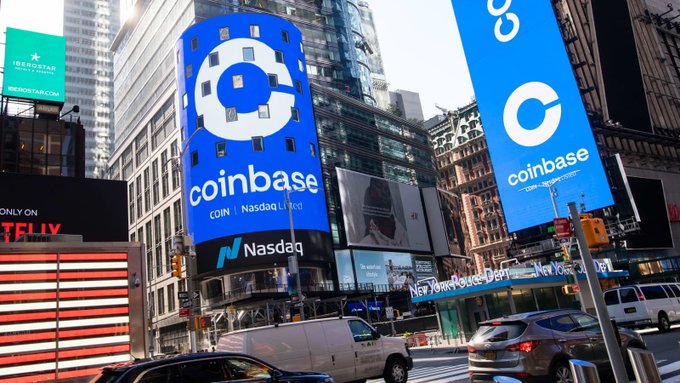 A street view in Times Square, New York, showing large blue digital billboards displaying the Coinbase logo and 'coinbase COIN | Nasdaq Listed' above a Nasdaq sign.
