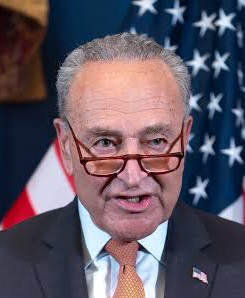A close-up portrait of Chuck Schumer speaking, wearing a dark suit with an orange tie and glasses, with an American flag in the background.