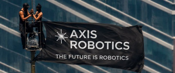 Two workers on a cherry picker hanging a large black banner that reads 'AXIS ROBOTICS - THE FUTURE IS ROBOTICS' against a glass building facade.