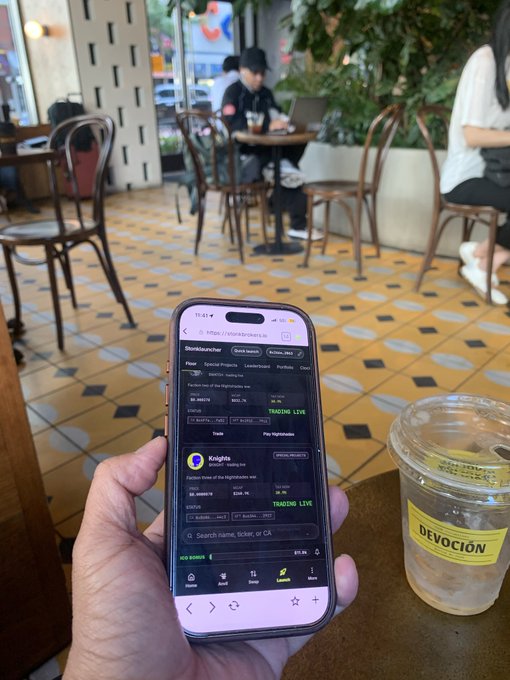 A hand holds a smartphone displaying a StoxDancer app showing trading data for 'Kugiga' with a 'TRENDING' badge, next to a clear plastic cup with a 'DEVOCIÓN' sticker, inside a cafe with yellow tiled floors.