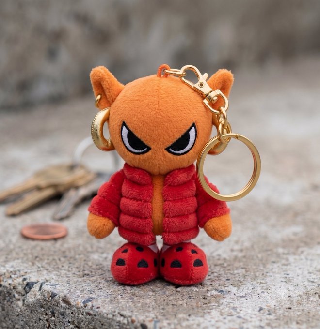 An orange plush cat character with an angry expression, wearing a red puffer jacket and red boots, is attached as a keychain to gold rings and traditional keys, echoing the post's appreciation for high-quality design and craftsmanship in the NFT space. The collectible figure showcases the creative artistry highlighted by the original poster, blending plush toy aesthetics with stylish character design.