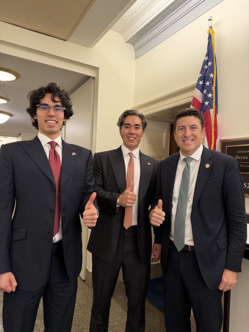 Three men in business suits smiling and giving thumbs up in a government building hallway, with an American flag and a "REPRESENTATIVE Bryan" door plaque visible behind them.