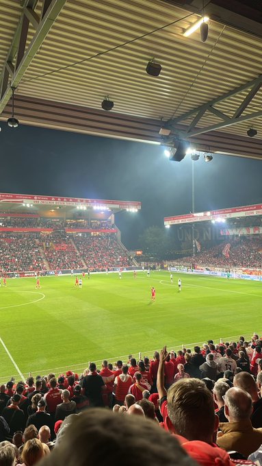 An evening view of a soccer match in a stadium shows a packed crowd in red watching the game under bright stadium lights, with 'UNION' visible on a banner.