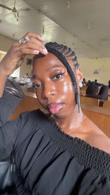 A selfie of a woman with braided cornrow hair and glossy makeup, wearing a black off-the-shoulder top. She poses in what appears to be a salon with a ceiling fan visible above.