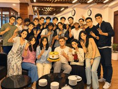 A group photo of about twenty young people gathered around a table with a birthday cake, all posing and gesturing toward the camera in what appears to be a restaurant setting.