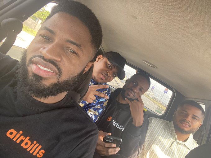 A selfie of four young men inside a car, with the person in the foreground wearing a black shirt with 'airbills' printed in orange.