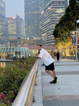 A man in a white shirt and black shorts stretches against a railing in an urban plaza, with modern skyscrapers and an arched glass structure visible in the background.