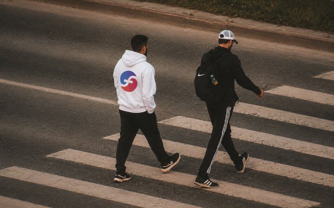 Two men walk across a zebra crosswalk on a street; one wears a white hoodie with a red and blue swirl logo on the back and black pants, while the other wears a black hoodie with a white Nike logo, a backpack, and a white cap.