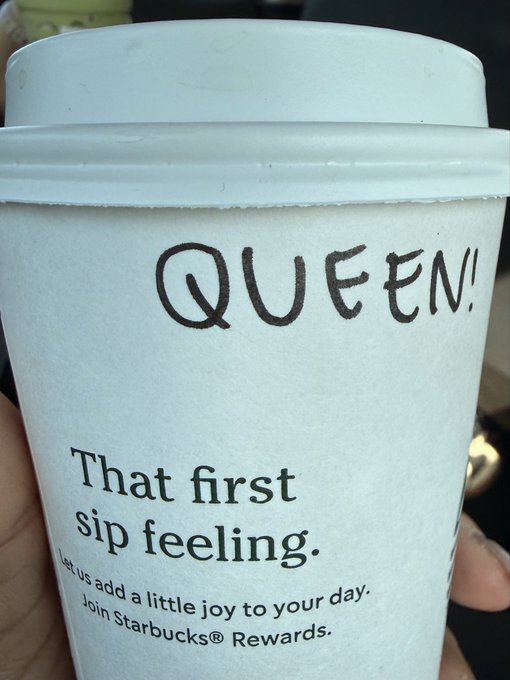 A Starbucks white paper coffee cup held in someone's hand with 'QUEEN!' handwritten in black marker on the lid, showing the cup's printed text 'That first sip feeling. Let us add a little joy to your day. Join Starbucks Rewards.'