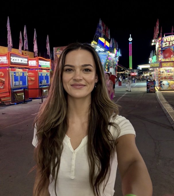 A woman with long brown hair in a white top takes a selfie at a brightly lit carnival or fairground at night, with colorful food stands visible behind her.
