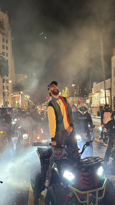A man stands confidently on an ATV at night during what appears to be a group ride, wearing a yellow, green, and red striped shirt with a crowd of motorcyclists behind him.