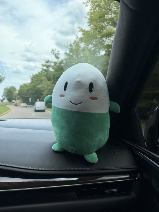 A green and white egg-shaped plush toy with a smiling face sits on the dashboard of a car, with trees and a road visible through the windshield.