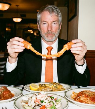 The image shows a man sitting at a table in front of plates filled with food. He is wearing a suit, tie, and holding a piece of food over his head as if he's about to take a bite or share it with someone else. The background reveals that they are likely having dinner together at a restaurant.