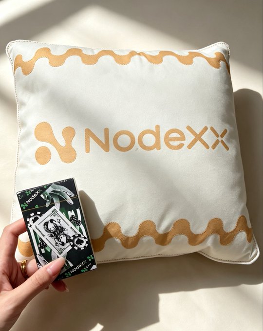 A hand holds a deck of NodeXX-branded playing cards in front of a cream-colored pillow with orange wavy trim and the "NodeXX" logo printed in the center, showcasing the branded merchandise set against a sunlit surface.
