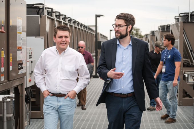 Two men in business casual clothing walking and conversing in an outdoor industrial setting, with other people visible in the background.