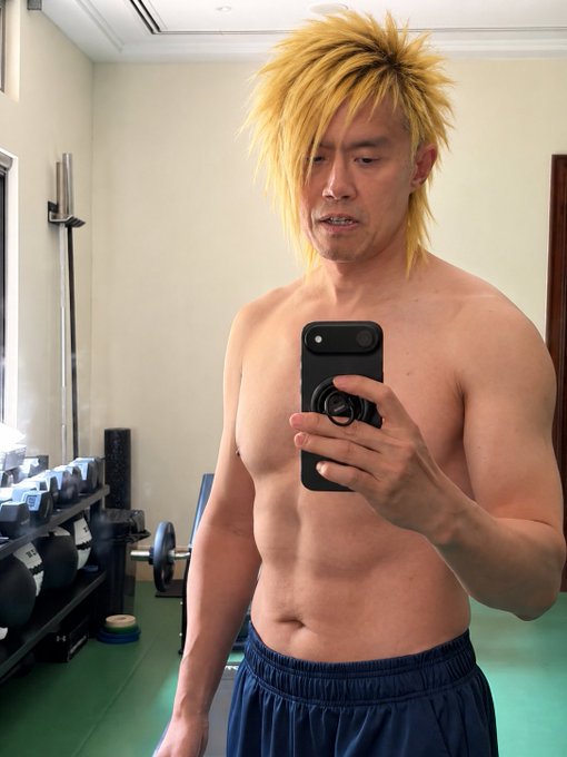 Mirror selfie of a shirtless, muscular man wearing a blonde spiky anime wig and blue shorts, holding a phone in a gym setting.