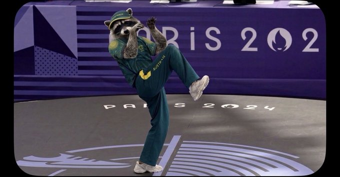 The Paris 2024 Olympics mascot, a Phrygian cap-wearing figure in green track pants with 'AUSTRALIA' on the leg, performing a breakdancing pose at an event venue.