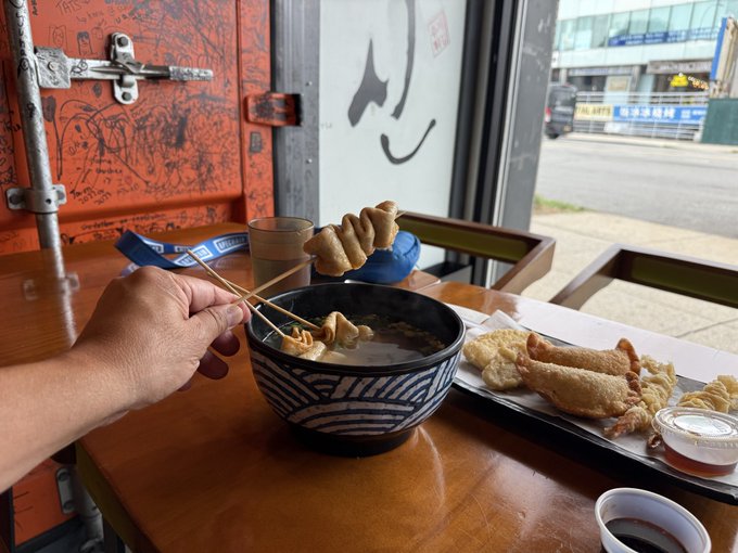 A hand using chopsticks to hold fried fish cake skewers over a bowl of noodle soup at a restaurant table, with a tray of additional fried seafood and dipping sauce nearby.