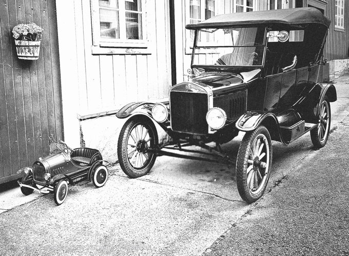 A black-and-white photograph showing a vintage Ford Model T-style car parked on a street next to a miniature toy version of the same vehicle, displayed in front of a wooden building with a hanging flower basket.