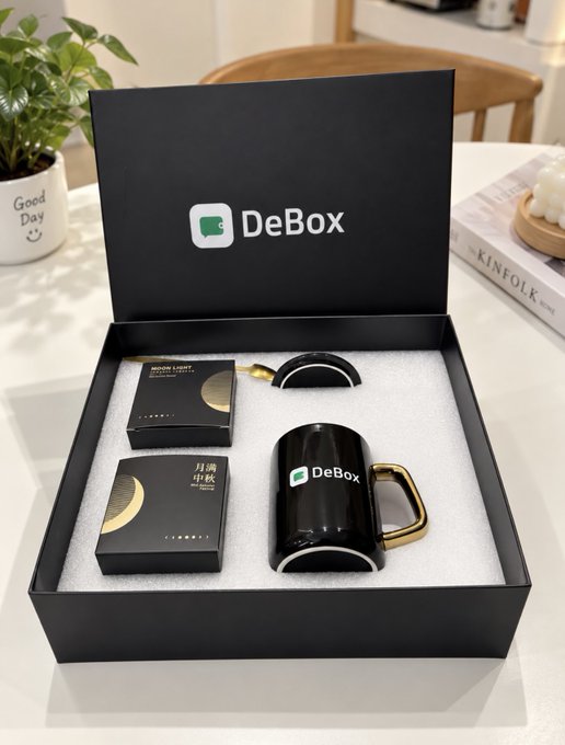 An open black gift box displays a DeBox branded mug set, including a black mug with gold handle, its lid, and two small black boxes featuring gold moon designs and Chinese text '月满中秋'.