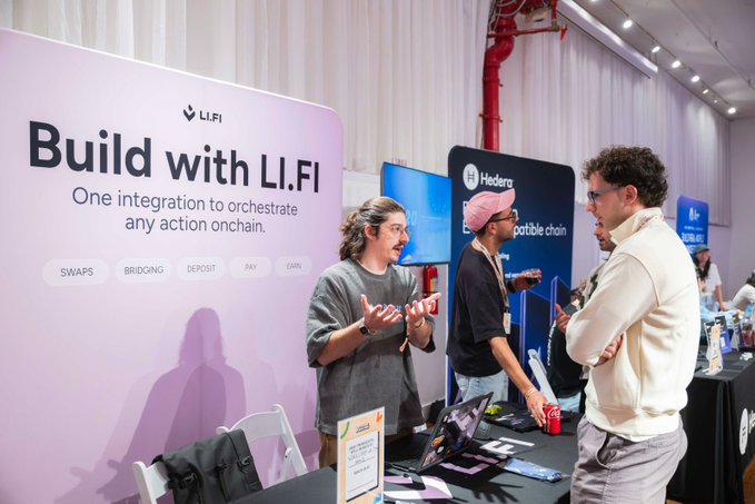 The image shows a group of people gathered around a table at an event. There are three individuals standing near the booth, engaged in conversation with each other. The booth they are standing by has a sign that says "Build With LI.FI". A man is wearing a pink hat and holding a cup of coffee while talking to another person. Another individual can be seen standing next to them.

The event appears to be focused on blockchain technology, as there is a display table with various items related to the topic. The attendees are likely discussing the potential applications and benefits of using LI.FI for cross-chain transactions or other blockchain projects.