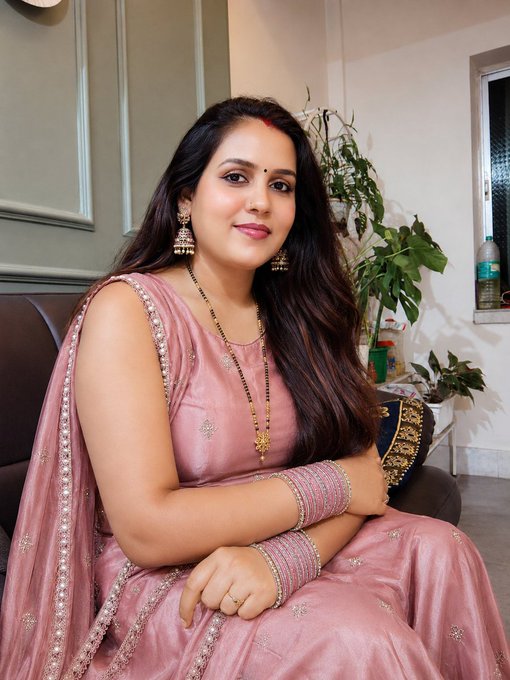 A woman wearing a pink traditional Indian sari with embroidered details, gold jewelry, and bangles poses indoors near a plant.