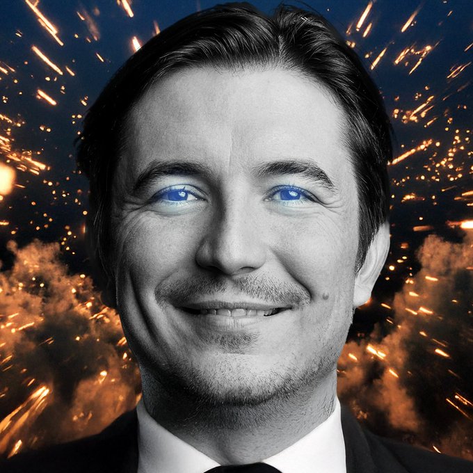 A black and white portrait of a smiling man with glowing blue eyes, set against a dramatic background of orange sparks and fire.