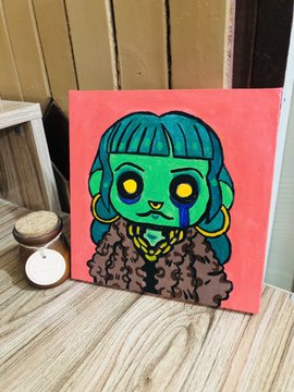A vibrant painting on a red canvas depicting a green-skinned figure with yellow eyes, blue hair, gold hoop earrings, and a leopard-print coat, displayed on a wooden surface next to a candle and jar.