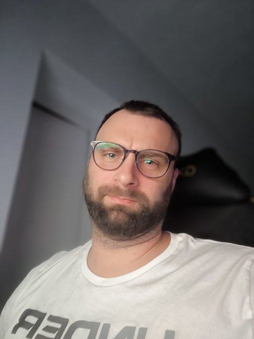 A selfie of a bearded man wearing clear-framed glasses and a white t-shirt, photographed against a light-colored wall.