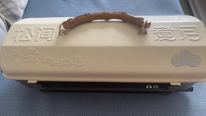 A heavy-duty cream-colored metal box with a decorative handle features embossed Chinese characters "松间月下" (Under the Moonlight Among Pines) along with floral engravings and a USDD token logo, sitting on a blue fabric surface.