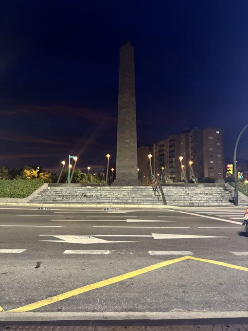 A tall, illuminated obelisk monument photographed at night with a wide pedestrian crosswalk in the foreground and apartment buildings visible in the background.
