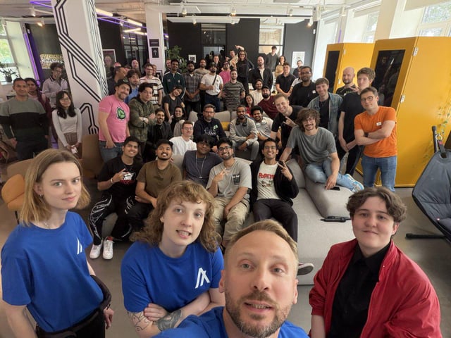 The image shows a large group of people gathered together, possibly for an event or meeting. They are standing around each other, with some individuals posing for a photo while others appear to be engaged in conversation. The room has a casual atmosphere, as evidenced by the presence of couches and chairs scattered throughout the space. 

The image also contains text that reads "The 36 hour Algorand hackathon in Berlin is still in full swing ... teams will be presenting their Agentic Commerce x402 projects today in front of a panel of judges." This suggests that there might have been an event or competition taking place, and the group of people present could be participants or organizers.