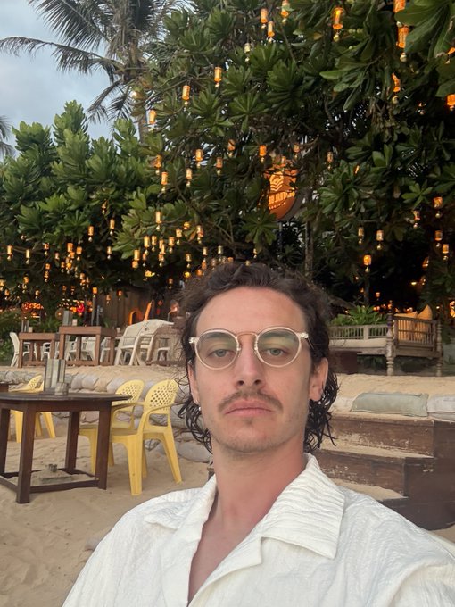 A young man with curly dark hair and round glasses takes a selfie at a beachside restaurant, wearing a white open-collar shirt with string lights hanging from trees in the background.