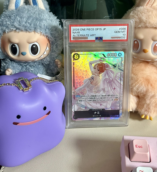 A graded PSA 10 Gem Mint 2026 One Piece OP15 Japanese alternate art card of Nami, displayed in its protective case alongside a purple Ditto Pokémon figure and Labubu plush toys.