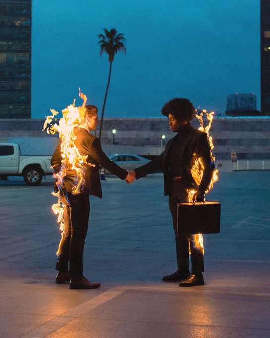 Two men in suits shake hands in a parking lot at dusk while both are engulfed in flames; one holds a briefcase, with a palm tree and city skyline in the background.