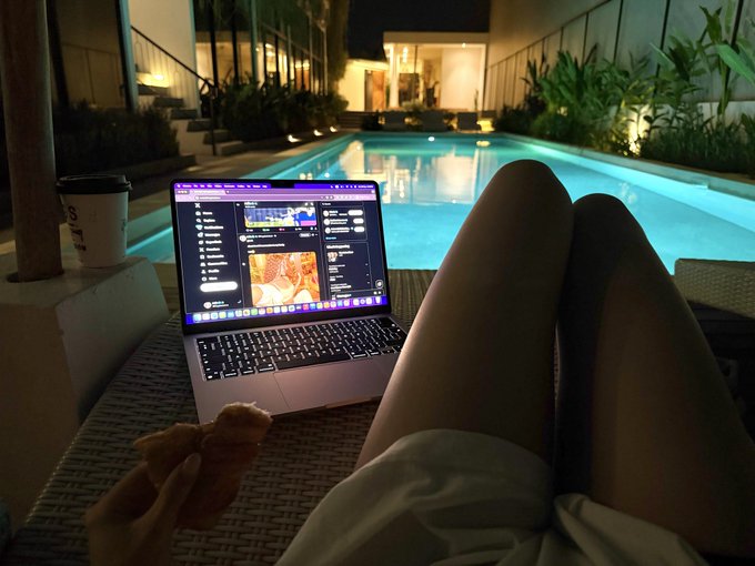 A person enjoys a peaceful Saturday night by the pool, lounging with a laptop (displaying a messaging app) and a pastry, accompanied by a takeaway coffee cup and a small teddy bear—capturing a quiet, content solo evening that feels more rewarding than a typical night out.