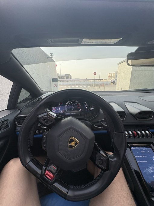 View from the driver's seat of a Lamborghini Huracán, with the iconic bull logo on the steering wheel, parked overlooking a cityscape—likely posted to represent an exotic Italian alternative to the post's repeated "GM" (General Motors) ticker.