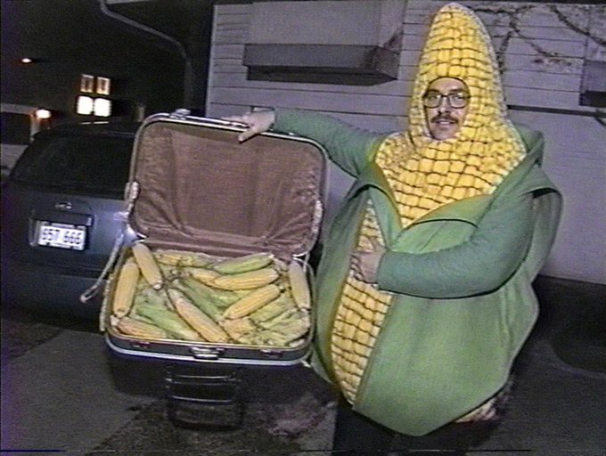 A man dressed in a corn cob costume with green husks stands next to an open suitcase filled with actual corn on the cob, posed next to a car at night.