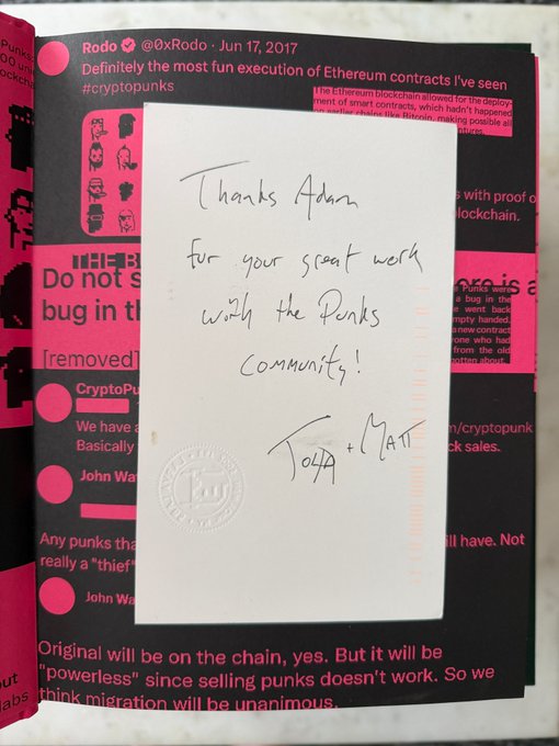 A handwritten note reading "Thanks Adam for your great work with the Punks community! John + Matt" resting on a book about CryptoPunks, with visible text about Ethereum blockchain and smart contracts by @0xRodo.