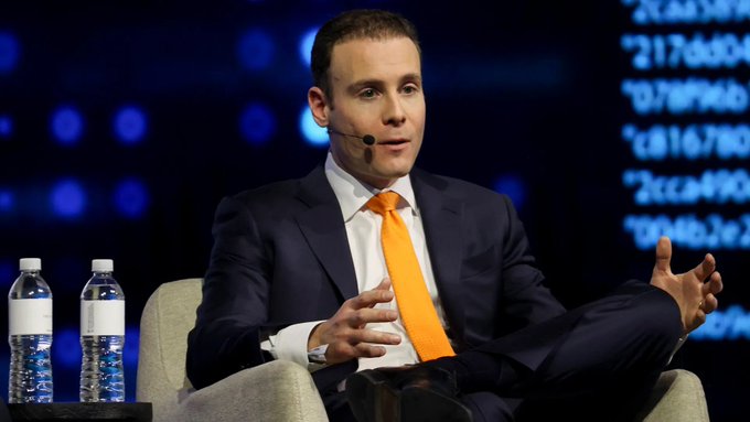 A man in a dark suit and orange tie speaks at a panel discussion, gesturing with his hands while seated beside water bottles, with hexadecimal code visible on a screen behind him. The image appears to relate to the post's coverage of crypto regulatory news, visually connecting the speaker to the announcement of the U.S. CFTC filing crypto asset rulemaking with the White House.