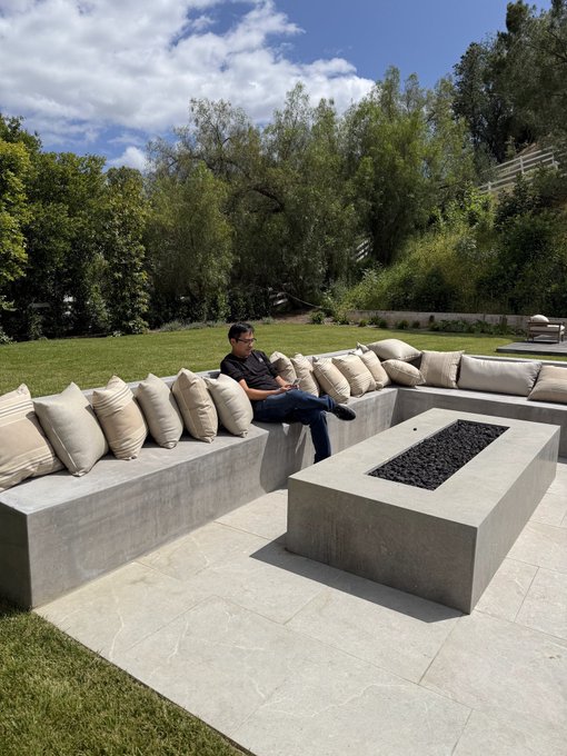 A man sits on a concrete outdoor sofa with cushions, using his phone next to a modern rectangular fire pit, surrounded by lush greenery.