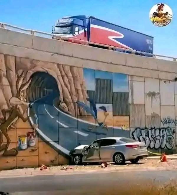 A silver car appears to have crashed into a wall painted with a mural depicting a road emerging from a tree tunnel, while a red, white, and blue truck drives on an overpass above.